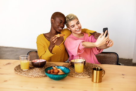 Beautiful young lesbian couple is doing selfie using a smart phone and smiling while having a breakfast at homeの写真素材