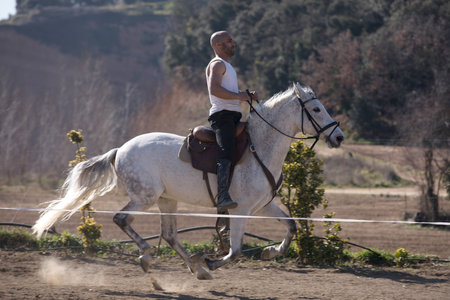 Young man in casual outfit riding white horse on sandy groundの写真素材