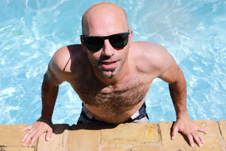 Portrait of a handsome man resting at the edge of a swimming pool wearing sunglasses. Summer vacation concept.の写真素材