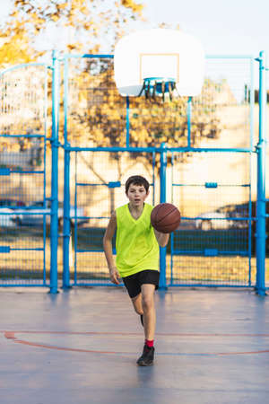 Cute boy in yellow shirt plays basketball on city playground. Active teen enjoying outdoor game with orange ball.の写真素材