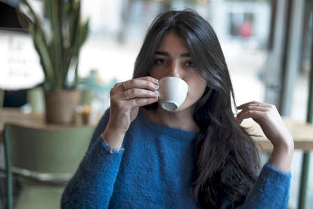 Young model look woman with long black hair is sitting in a coffee shop during the brunch. Latin female is looking down while sitting at the table in the cafe.の写真素材