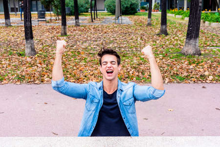 Close up portrait of a young man with arms raised in celebration outdoors in a public parkの写真素材