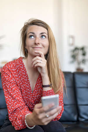 Beautiful Caucasian woman hold cellphone in hand and sitting on sofa while looking away thoughtfully