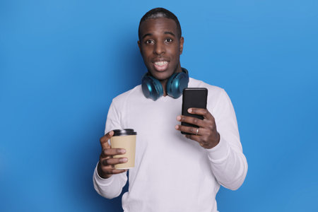 African man on blue background holding coffee to take away and a mobileの写真素材