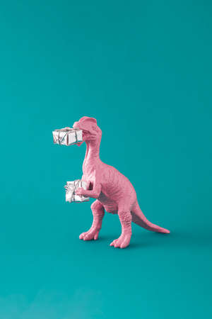 Pink painted dinosaur toy holding gifts for Christmas or New Year. Minimal winter holidays idea on green blue background. Creative composition.の写真素材