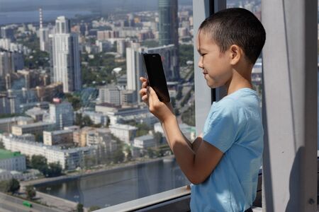 A Muslim boy uses a smarphone against the background of a panoramic window. Blurred view of the city of Yekaterinburg from a high-rise building.の写真素材