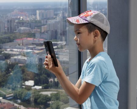 A Muslim boy uses a smarphone against background of a panoramic window. Blurred view of city of Yekaterinburg from a high-rise building.の写真素材