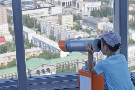 The boy looks into a telescope from a panoramic window of a high-rise building on the city of Yekaterinburg, Russiaの写真素材