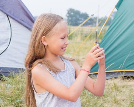 Cute small girl with long blond hair takes a picture on a smartphone on a background of tents.の写真素材