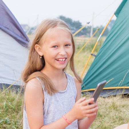 Cute small girl with long blond hair takes a picture on a smartphone and smile on a background of tents.の写真素材