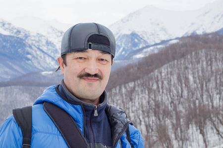 A tourist in a cap and blue jacket is looking at the camera and smiling. Close-up. Background - snow-capped mountains.の写真素材