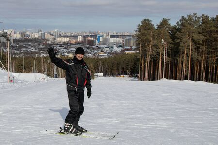 A man stands skiing looking at the camera and smiling. He raised his hand in greeting. In the background is the ski slope and the outskirts of the city of Yekaterinburg.の写真素材