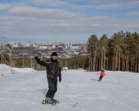 A man stands skiing, looking at the camera and smiling. He raised his hand in greeting. In the background is the ski slope and the surroundings of the city of Yekaterinburg.の写真素材