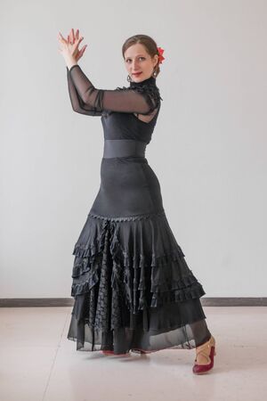 Portrait of beautiful girl in a long black dress and a red rose in her hair dancing flamenco on a gray wall background.の写真素材