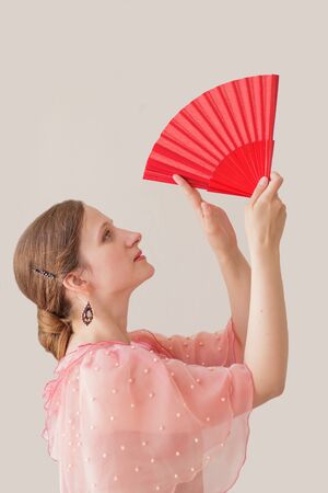 Portrait of a beautiful young woman in a beige and pink dress on a beige background. She is dancing flamenco. Hands with a red fan raised up.の写真素材