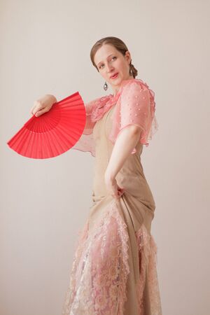 Portrait of a beautiful young woman in a beige and pink dress on a beige background. She is dancing flamenco with a red fan.の写真素材