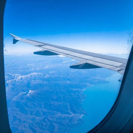 View of the wing of an airplane flying over the mountains and the sea through the porthole window.