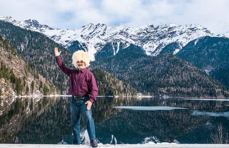 An Asian man in a traditional Caucasian white sheepskin hat, in a red shirt and jeans raised his hand in greeting as a backdrop of a beautiful mountain landscape and Lake Ritsa, Abkhazia.の写真素材