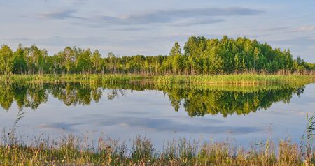 Beautiful summer countryside landscape. On the smooth water surface of the lake reflected green trees and grass, a blue cloudy sky. Banner. Siberia, Russiaの写真素材