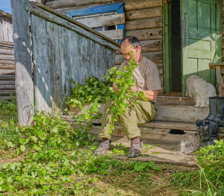Senior Asian man ties birch brooms for a bath sitting on a wooden porch of an old country house in a Siberian village, Russiaの写真素材