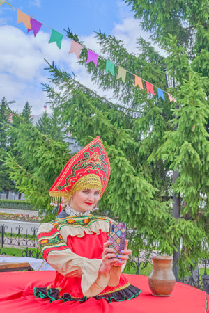 Aromashevo, Russia - 27 June, 2020: Young woman in kokoshnik in stylized red Russian folk clothes uses a smartphone for selfies at a street fair against the backdrop of a large green spruce.のeditorial素材