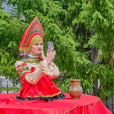 Aromashevo, Russia - 27 June, 2020: Young woman in kokoshnik in stylized red Russian folk clothes uses a smartphone at a street fair against the backdrop of a large green spruce.のeditorial素材