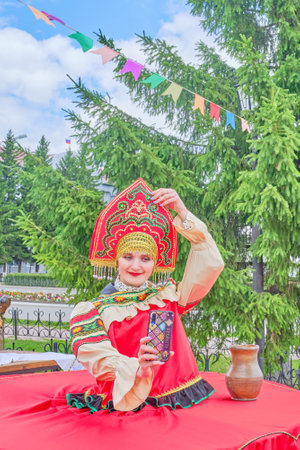 Aromashevo, Russia - 27 June, 2020: Young woman in kokoshnik in stylized red Russian folk clothes uses a smartphone for selfies at a street fair against the backdrop of a large green spruce.のeditorial素材
