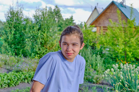 Portrait of a shyly smiling teenager in a blue t-shirt on the background of a vegetable garden and a country house.の写真素材
