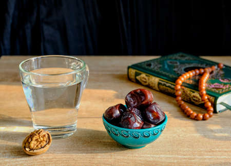 Walnut, turquoise piala with traditional oriental pattern with dates, glass of clear water, stone rosary and green and gilded book of Quran on table during sunset First meal, Muslim fast Eid al, Urazaの写真素材