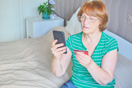 Senior woman feeling pain sitting on bed at home in casual clothes. In one hand she holds a pill, about to take it, in the other a cup of water.の写真素材