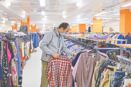 Mature man in mask choosing clothes at Second hand store. Conscious consumption, a new normal conceptの写真素材