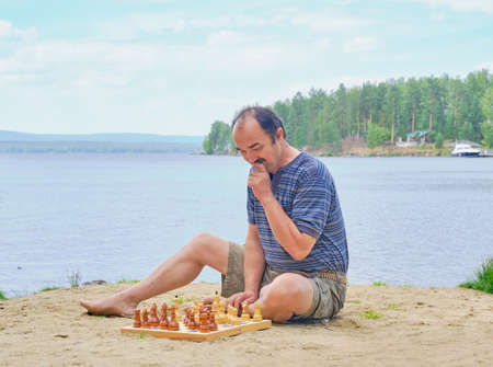 Senior asian man pondering a move in a game of chess and sitting on the beach near the lake. Digital detox conceptの写真素材