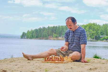 Senior asian man pondering a move in a game of chess and sitting on the beach near the lake. Digital detox conceptの写真素材