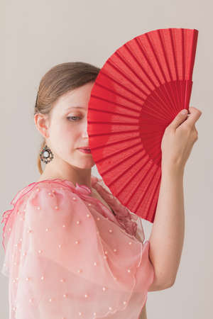 Portrait of a beautiful caucasian young woman in a pink dress covering half of her face with a red fan, on a beige backgroundの写真素材