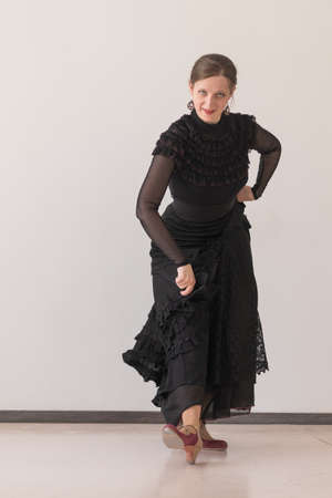 Portrait of beautiful young woman in a long black dress dancing flamenco and looking at the camera on a beige wall background. The foot is placed on the heelの写真素材
