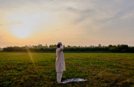 A Muslim senior man wearing an embroidered skullcap and white traditional clothes prays at sunset in a fieldの写真素材