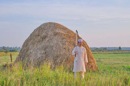 An senior Muslim man in a skullcap and traditional clothes leaves with a hand scythe from the hay field at sunset. Country life conceptの写真素材