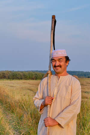 Portrait of smiling senior muslim man in an embroidered skullcap and white traditional clothes with hand-scythe in a hayfield. Looking at the camera. Country life conceptの写真素材