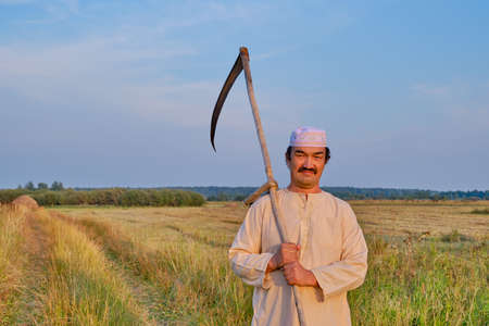 Portrait of senior asian man in an embroidered skullcap and white traditional clothes with hand-scythe in a hayfield. Looking at the camera. Country life conceptの写真素材