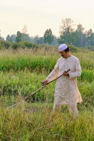 An senior asian man in an embroidered skullcap and white traditional clothes mows hand-scythe grass in a hayfield. Country life conceptの写真素材