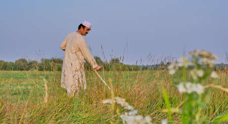 An senior asian man in an embroidered skullcap and white traditional Clothes mows hand-scythe grass in a hayfield. Country life conceptの写真素材