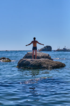 A male sunbathes in the sun with his arms outstretched standing on a stone among the sea. On a nearby stone stands a cormorant with open wingsのeditorial素材