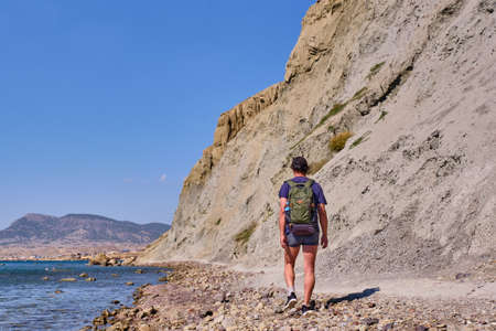 A tourist with a backpack walks along the path of the rocky coast of Cape Meganom on the Black Sea,...