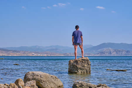 Senior asian man in shorts, t-shirt and baseball cap, holding in his hand in face mask, stands alone on a stone in a sea bay over a background of mountainsの写真素材