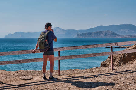 A tourist with a backpack standing near a wooden fence on the sea coast. He thinking about the route of the trip.の写真素材