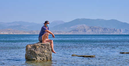 Senior asian man in face mask, shorts, t-shirt and baseball cap sits alone on a stone in a sea bay against a background of mountains Bannerの写真素材
