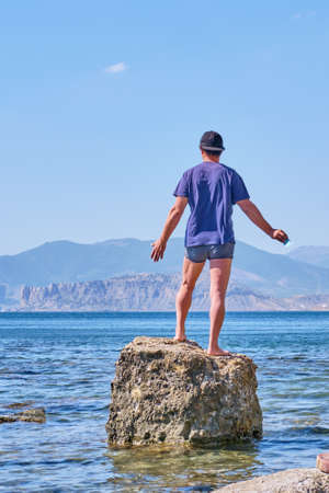 Senior asian man in shorts, t-shirt and baseball cap, holding in his hand face mask, stands alone on a stone in a sea bay over a background of mountainsの写真素材