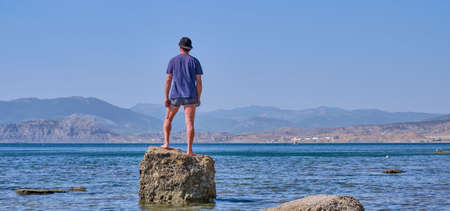 Senior asian man in shorts, t-shirt and baseball cap, holding in his hand in face mask, stands alone on a stone in a sea bay over a background of mountains. Bannerの写真素材