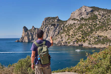 Black Sea coast, Crimean peninsula. An Asian senior man with a backpack takes photos on a smartphone over the background of the Blue Bay on tourist route Golitsyn trail. Botanical reserve New Worldの写真素材