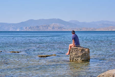 Senior man in shorts and baseball cap sits alone on a stone in a sea bay over a background of mountainsの写真素材